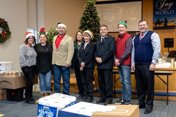 eight people stand in a room with a Christmas tree in the background and donated items around them.