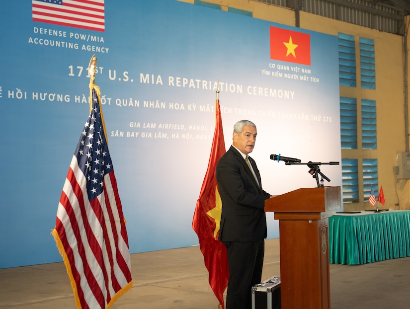 A man in a suit speaks into a microphone while standing in front of a lectern. The American and Vietnamese flags are displayed behind him.