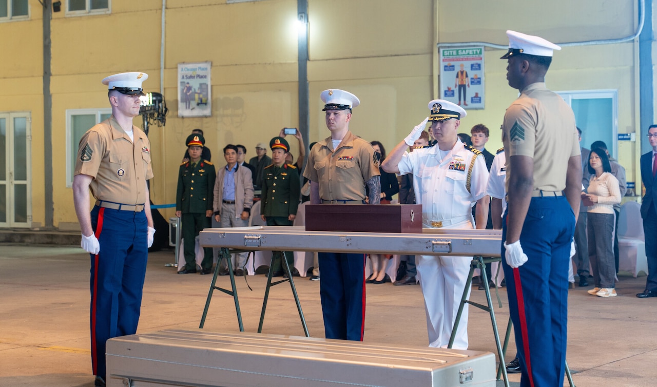 A man in a military dress uniform salutes a box filled with human remains as three service members in dress uniforms stand at attention around the box. There are people in military dress uniforms and civilian attire standing in the background.