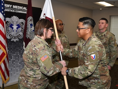 Capt. Justine Juan, right, commander of U.S. Army Medical Logistics Command’s Headquarters and Headquarters Detachment, passes the unit guidon to incoming detachment sergeant, Sgt. 1st Class Brandi Brown, during a change of responsibility ceremony Dec. 12, 2025, at Fort Detrick, Md. Also pictured are Sgt. 1st Class Aaron Taylor, background center, and outgoing detachment sergeant, Master Sgt. William Harbeson.