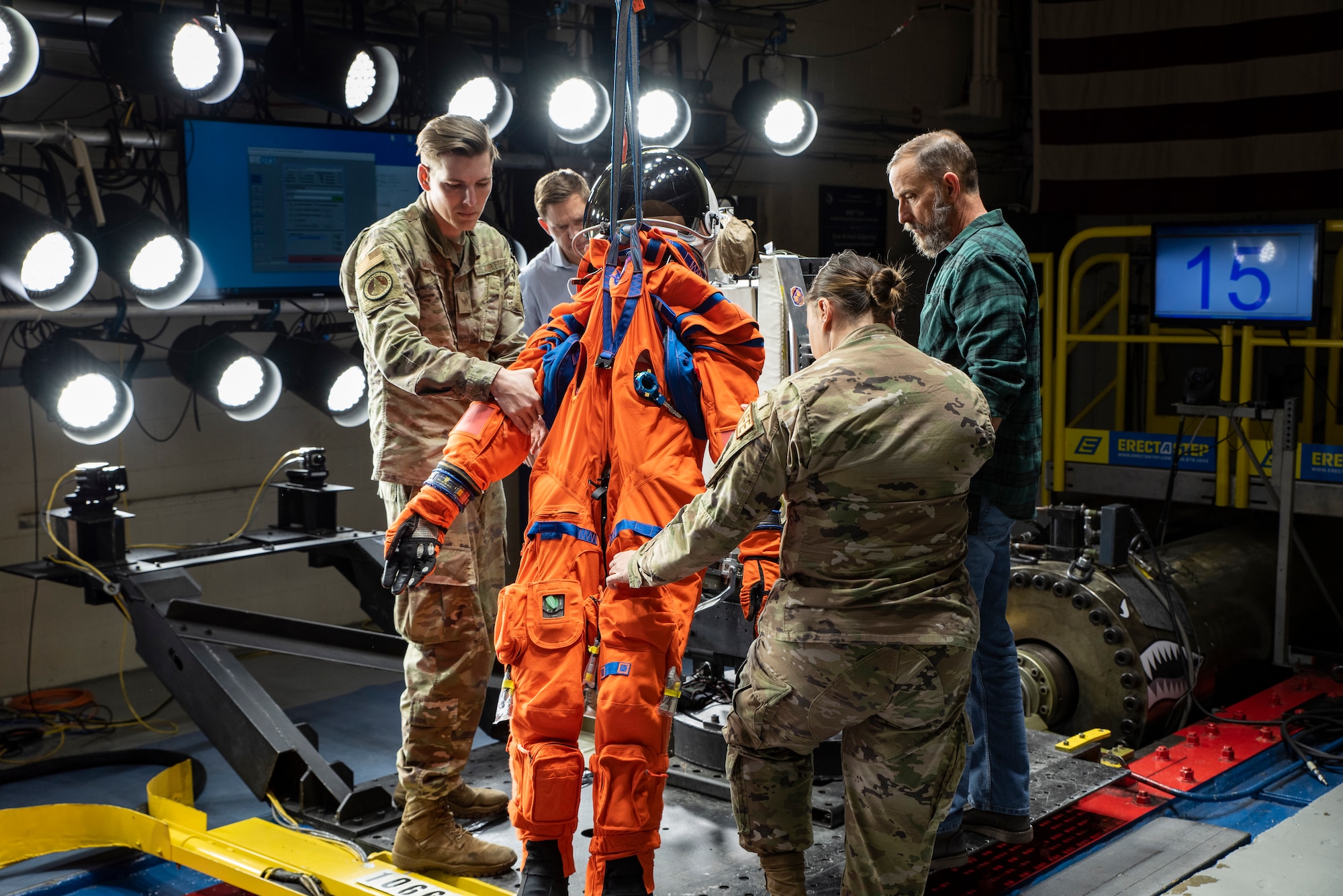 In a high-tech lab, four individuals, two civilian and two uniformed military, tinker with an orange spacesuit propped up by a harness