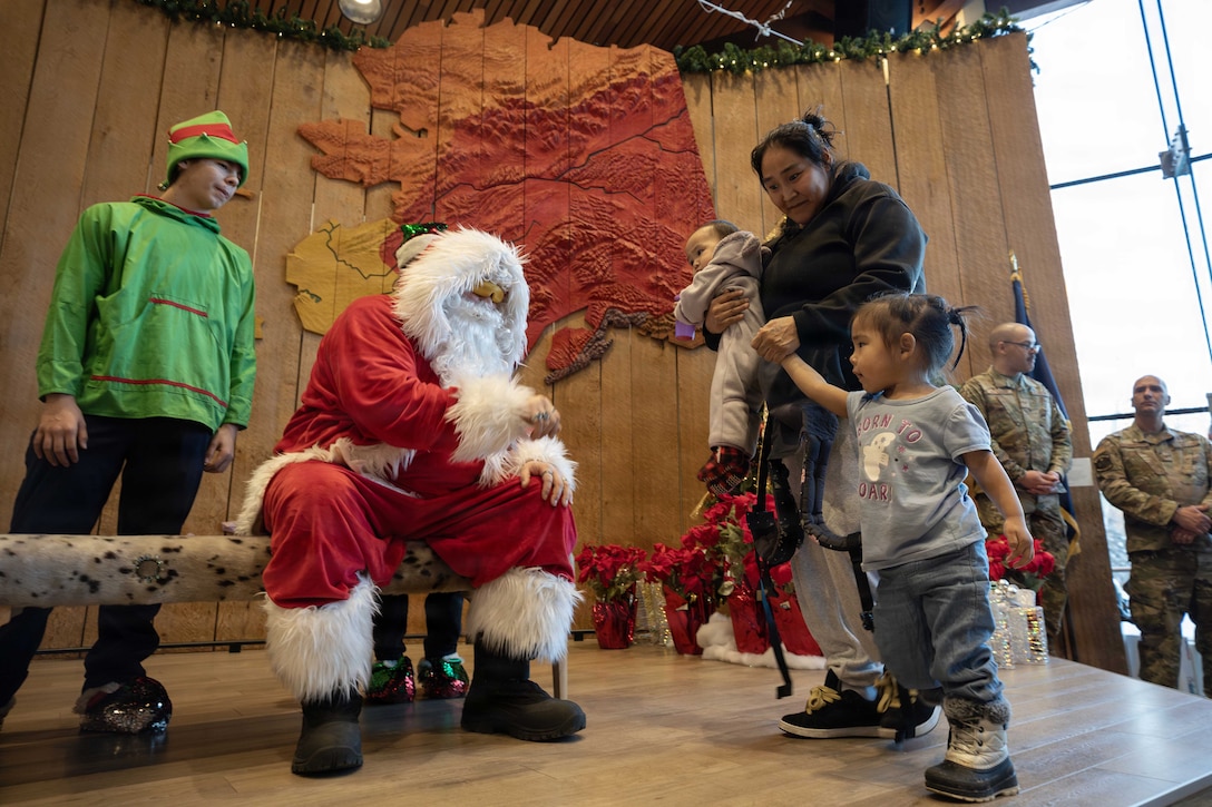 A parent holds a baby in one hand and a toddler's hand in the other as they approach a seated Santa Claus and standing elf in a dimly lit room.