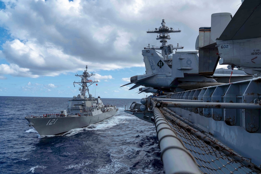 Two ships connected by a fuel hose transit a body of water during the day.