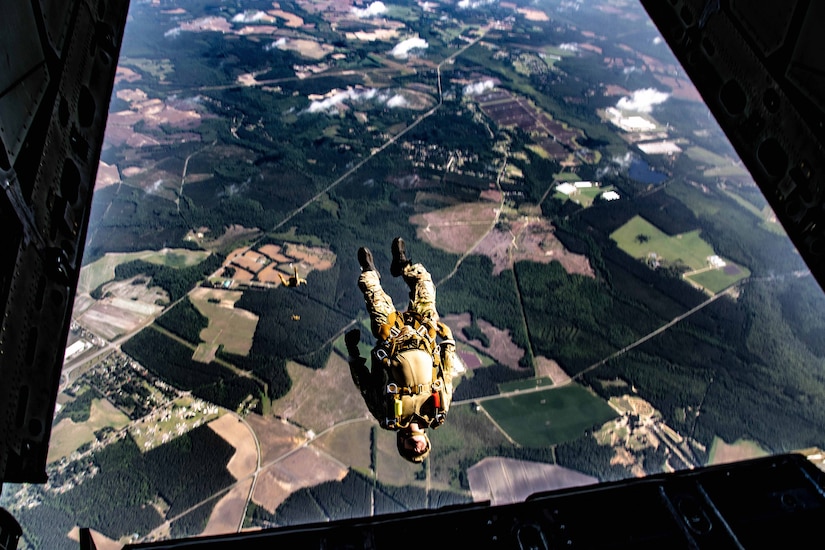 A man in a camouflage military uniform jumps from the back of an aircraft into the air thousands of feet above the ground.