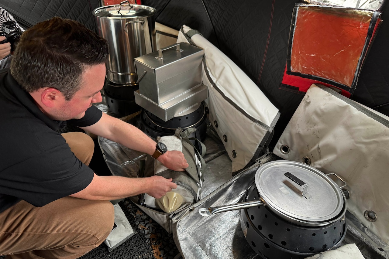 A man in business casual attire crouches down to open a container beside large bags that contain pots and other metal items.