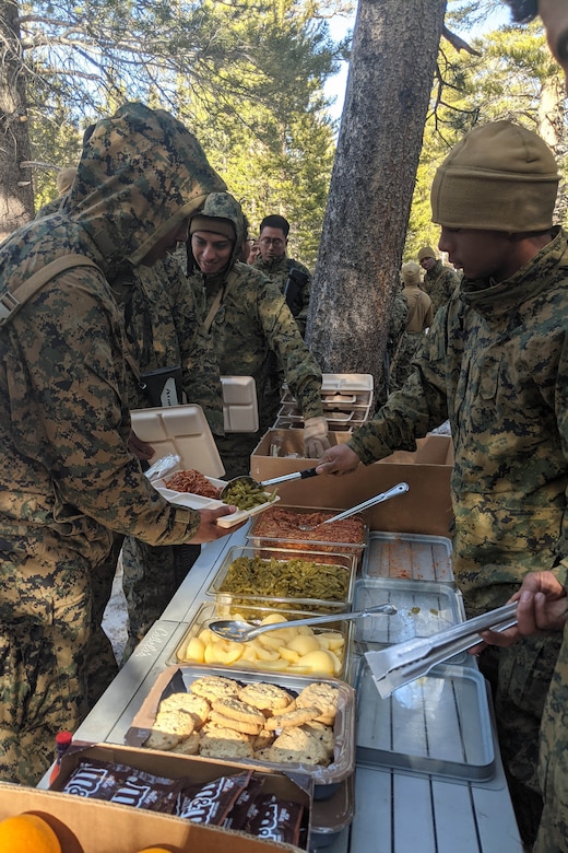 A man in a military camouflage uniform serves food to another man in similar attire as others wait in line behind him to be served.