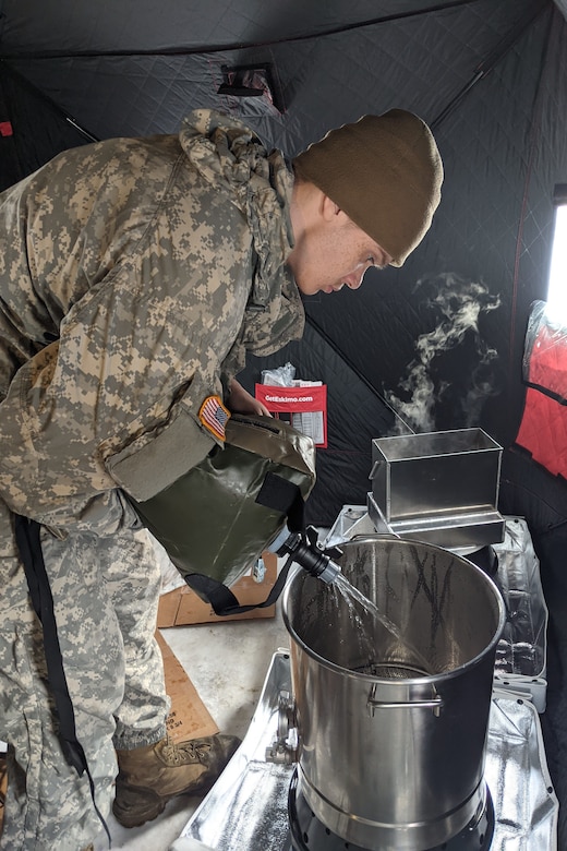 A man wearing a camouflage military uniform pours water from a large pouch into a large pot while standing in a tent.