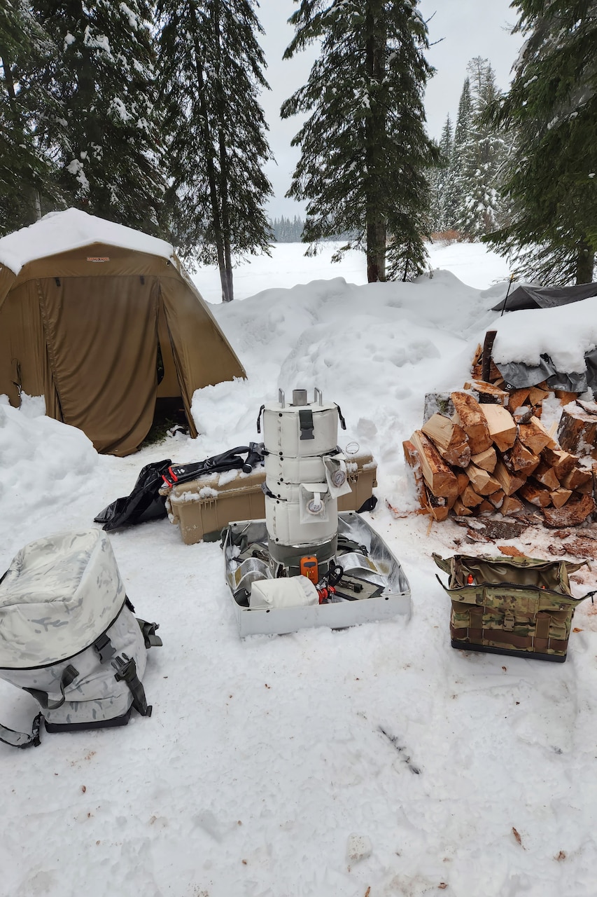 A snowy campsite includes a stove, a tent, bags, bins and a pile of logs.