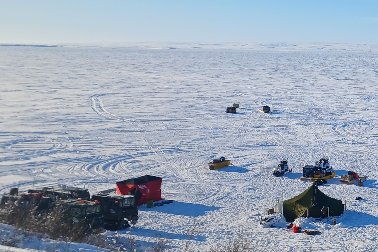 On a vast landscape of white snow, a few vehicles and two tents sit.