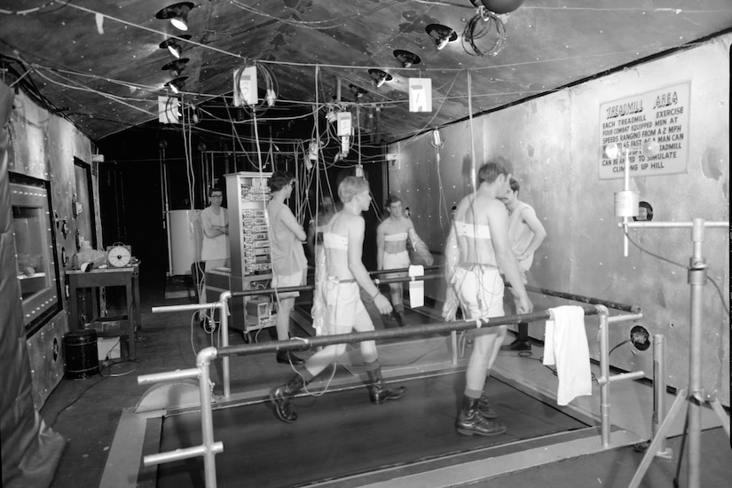 Three men in shorts and boots walk on large treadmills inside a massive chamber filled with wires and gauges.