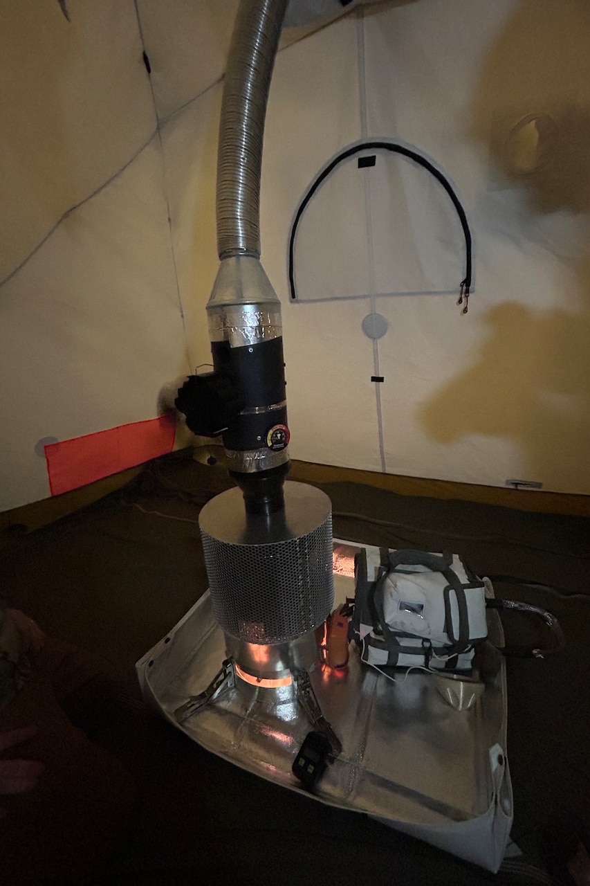 Inside a tent, a small stove sits in a basin. A metal tube vents toxins outside of the tent from the stove.