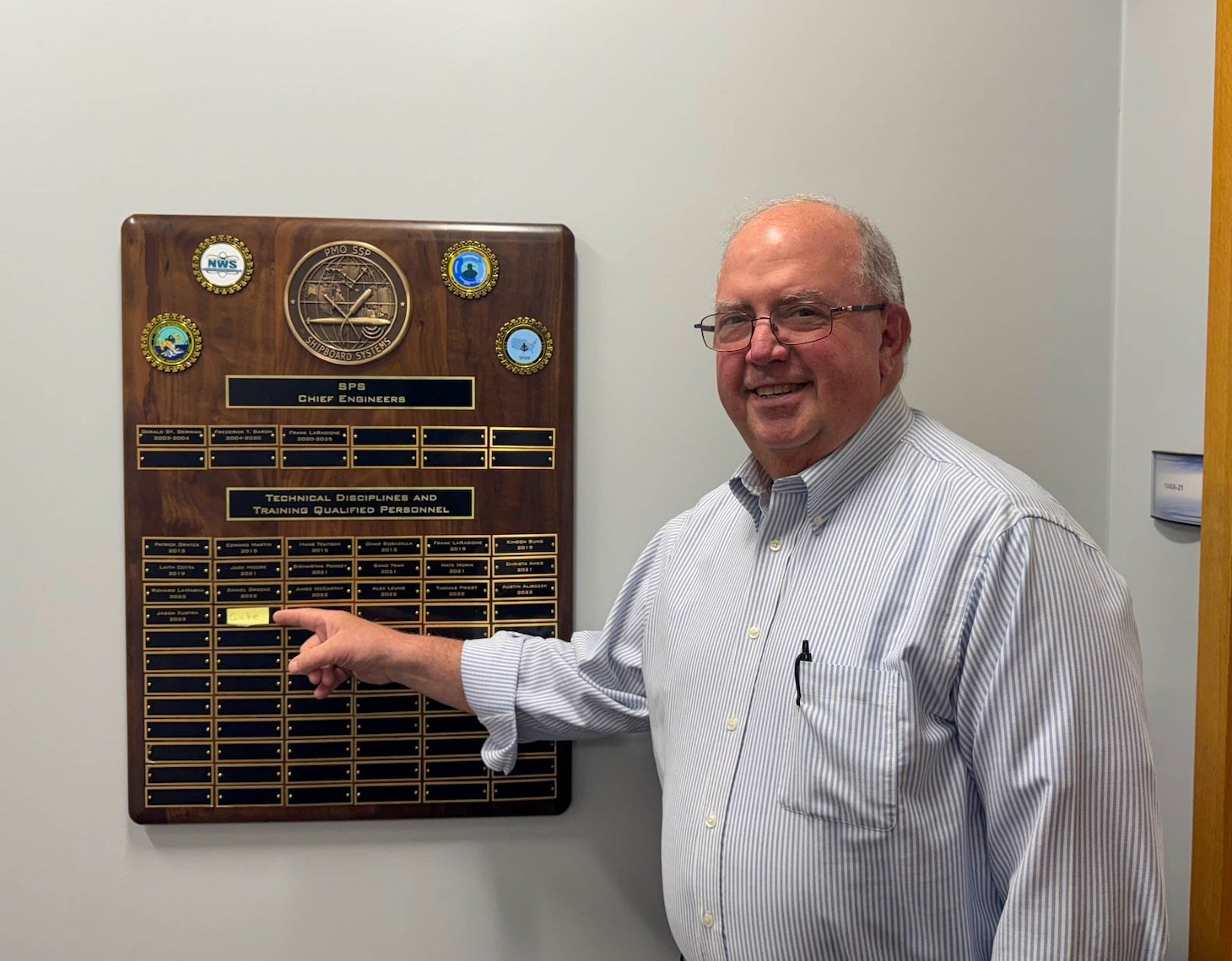 Pittsfield, Mass. - Frank LaRagione, chief engineer at PMOSPS (retired), points to a new entry placeholder on a plaque in Pittsfield, Mass., in Spring 2025. The plaque recognizes members of the PMOSPS technical workforce that have completed the Technical Discipline Training and Qualification Program (TDTQP). TDTQP provides a fundamental understanding of the technical policies, processes, and acquisition management tools that underpin the nation’s Trident II D5 strategic weapons system (SWS), nuclear-armed sea launched cruise missile (SLCM-N), and the non-nuclear hypersonic conventional prompt strike system (CPS).