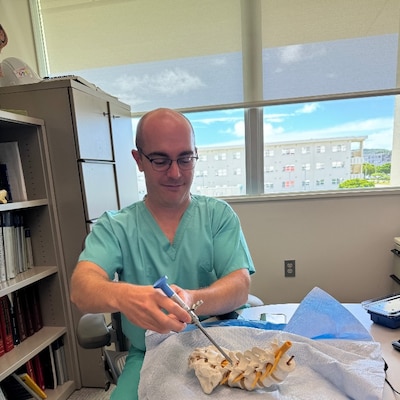 251217-N-TW242-1003 Okinawa, Japan Dr. Lawless explains the surgical procedure in his office with a spine visual aid at USNHO (U.S. Navy photo by Public Affairs Officer Trey Savitz)