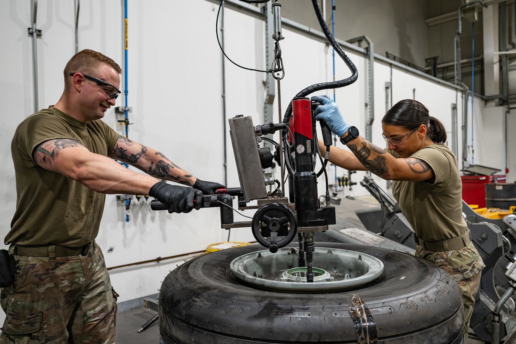 Two Airmen stand on either side of a tire. The Airman on the left holds two handles of a machine while the other holds it at the top.
