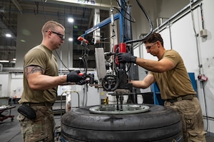 Two Airmen stand on either side of a large tire wearing protective equipment while operating machinery hovering over the tire