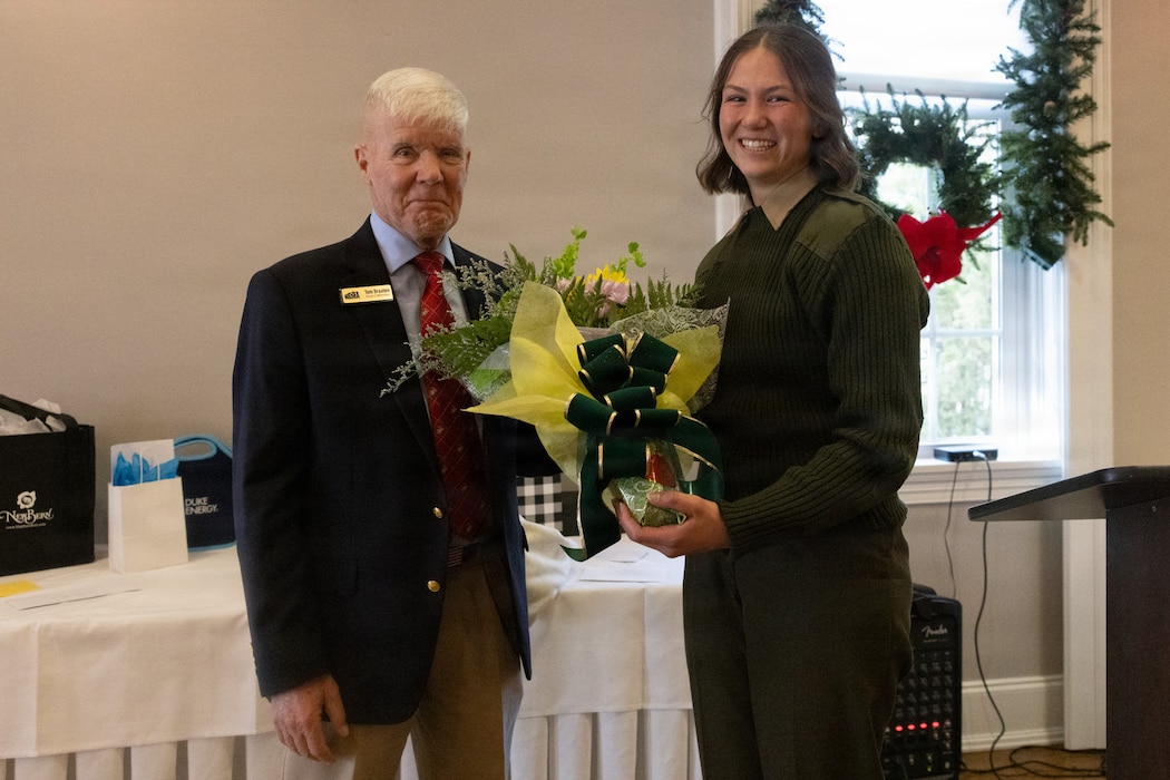 Retired U.S. Marine Corps Maj. Gen. Tom Braaten, left, presents Cpl. Mya Seymour, a combat graphics specialist with 2nd Marine Aircraft Wing, with flowers during a Service Person of the Quarter ceremony at the New Bern Golf and Country Club, New Bern, North Carolina, Dec. 11, 2025. The New Bern Military Alliance honored Seymour for a commitment to service that extends far beyond her duties, recognizing her selfless contributions to local non-profits and her dedication to mentoring children. (U.S. Marine Corps photo by Lance Cpl. Emma Powell)