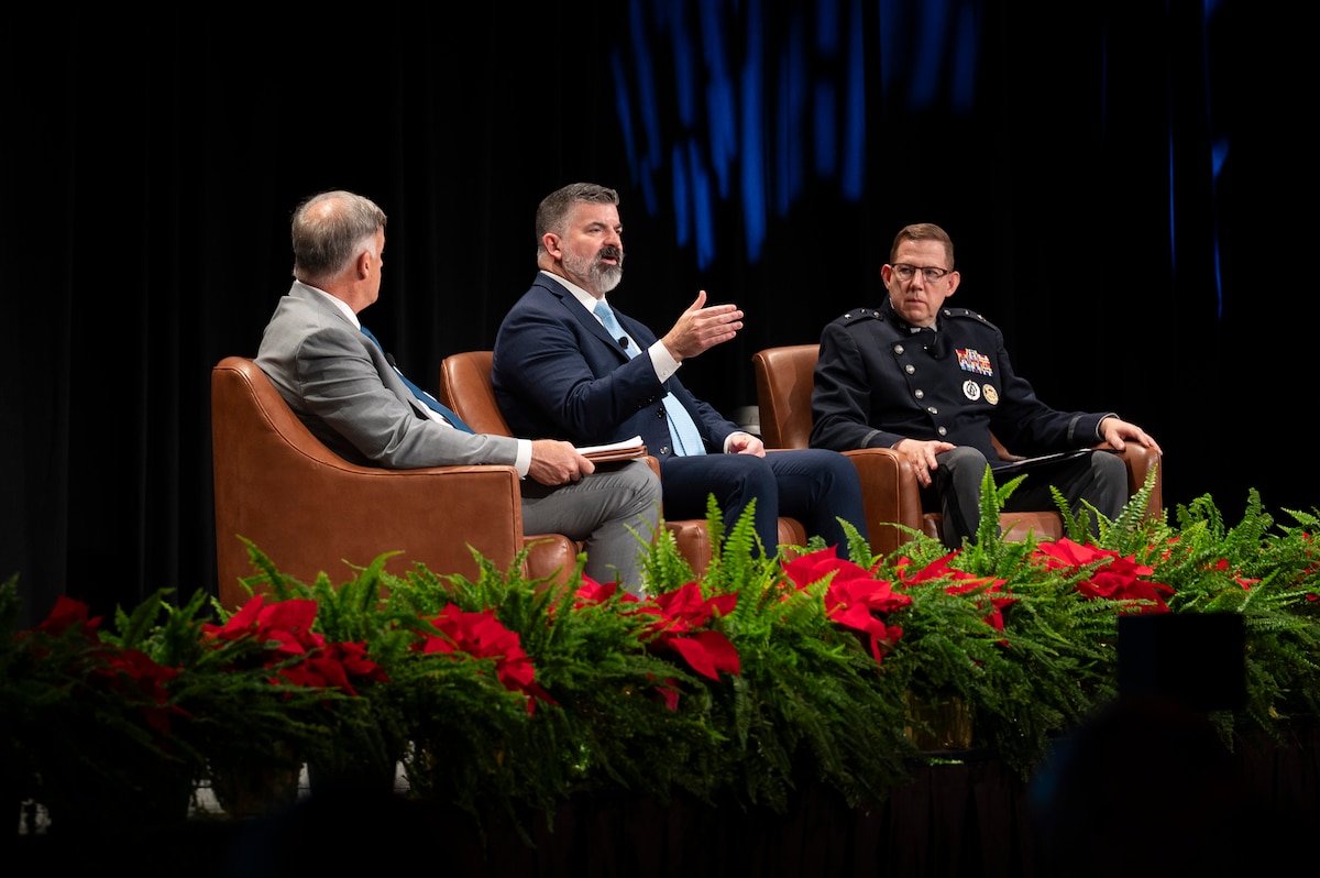 Three men sit in chairs on stage, speaking. Red flowers are in front of them.