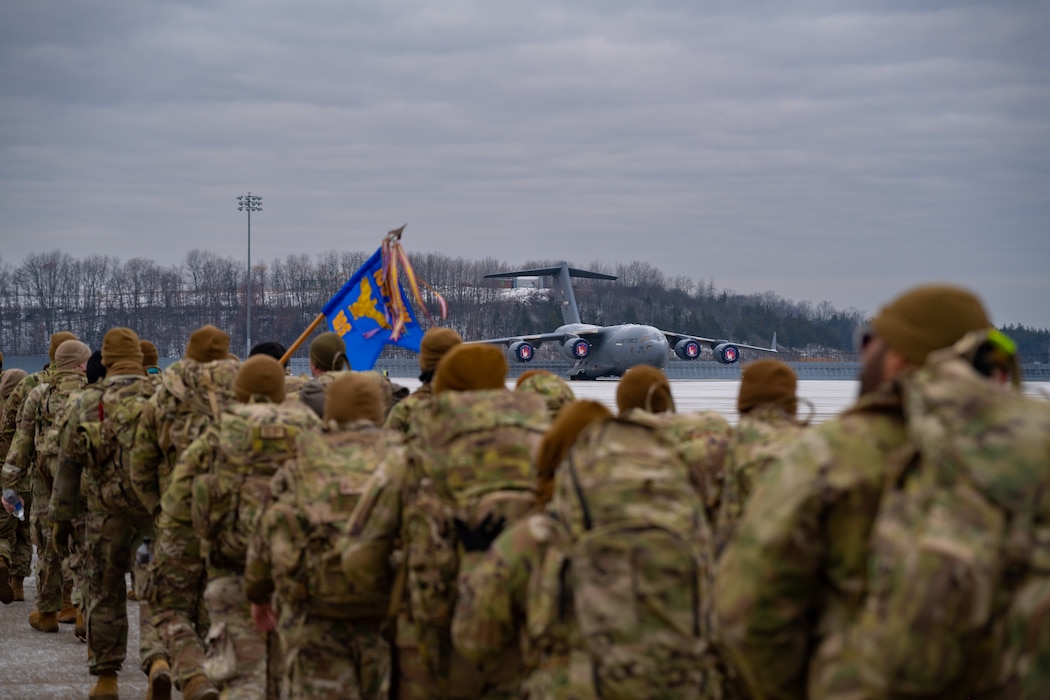 U.S. Airmen in winter uniforms march across a grey-skyed flightline with U.S. aircraft in the background.