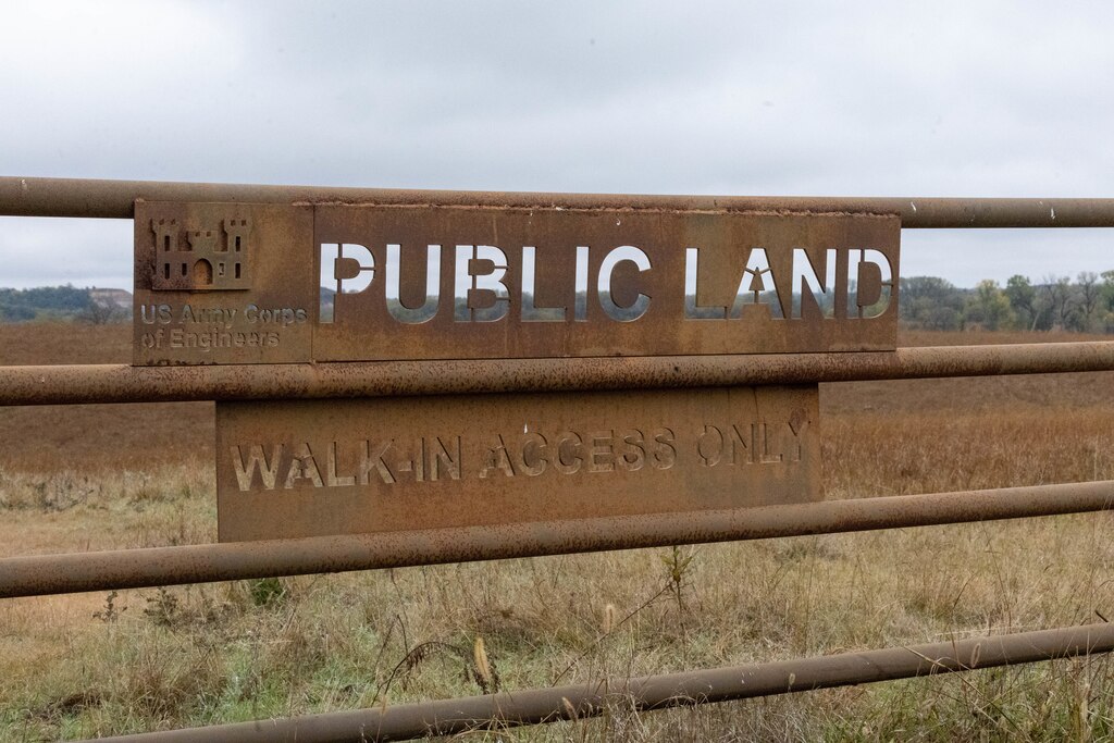 A brown rusted metal sign that says Public Land, Walk In Access Only with brown vegetation, trees, and grey sky in the background.