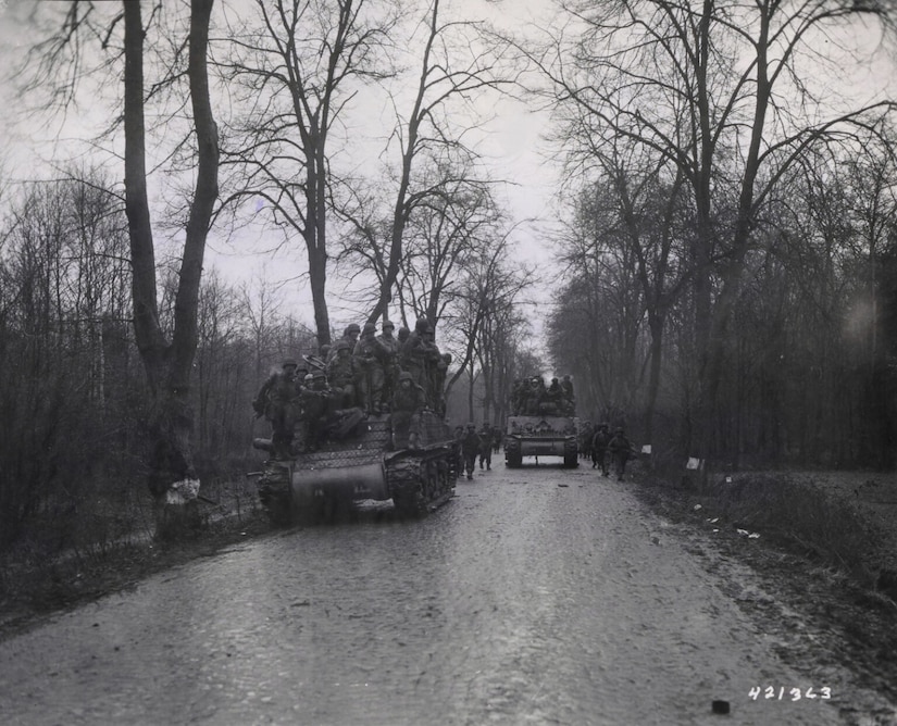 Several men in military uniforms ride on the outside of two tanks as they move along a paved road lined with trees. Several other soldiers in similar attire march behind the tanks.