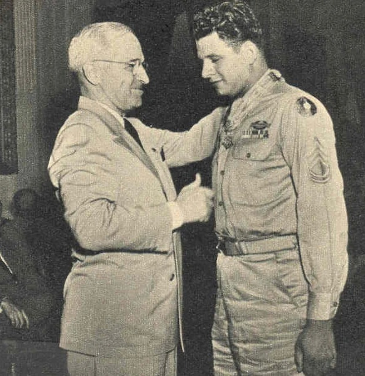 An older man in a suit puts his hand on the shoulder of a younger man in a military dress uniform.