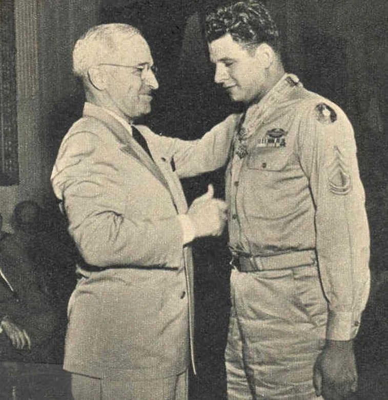 An older man in a suit puts his hand on the shoulder of a younger man in a military dress uniform.