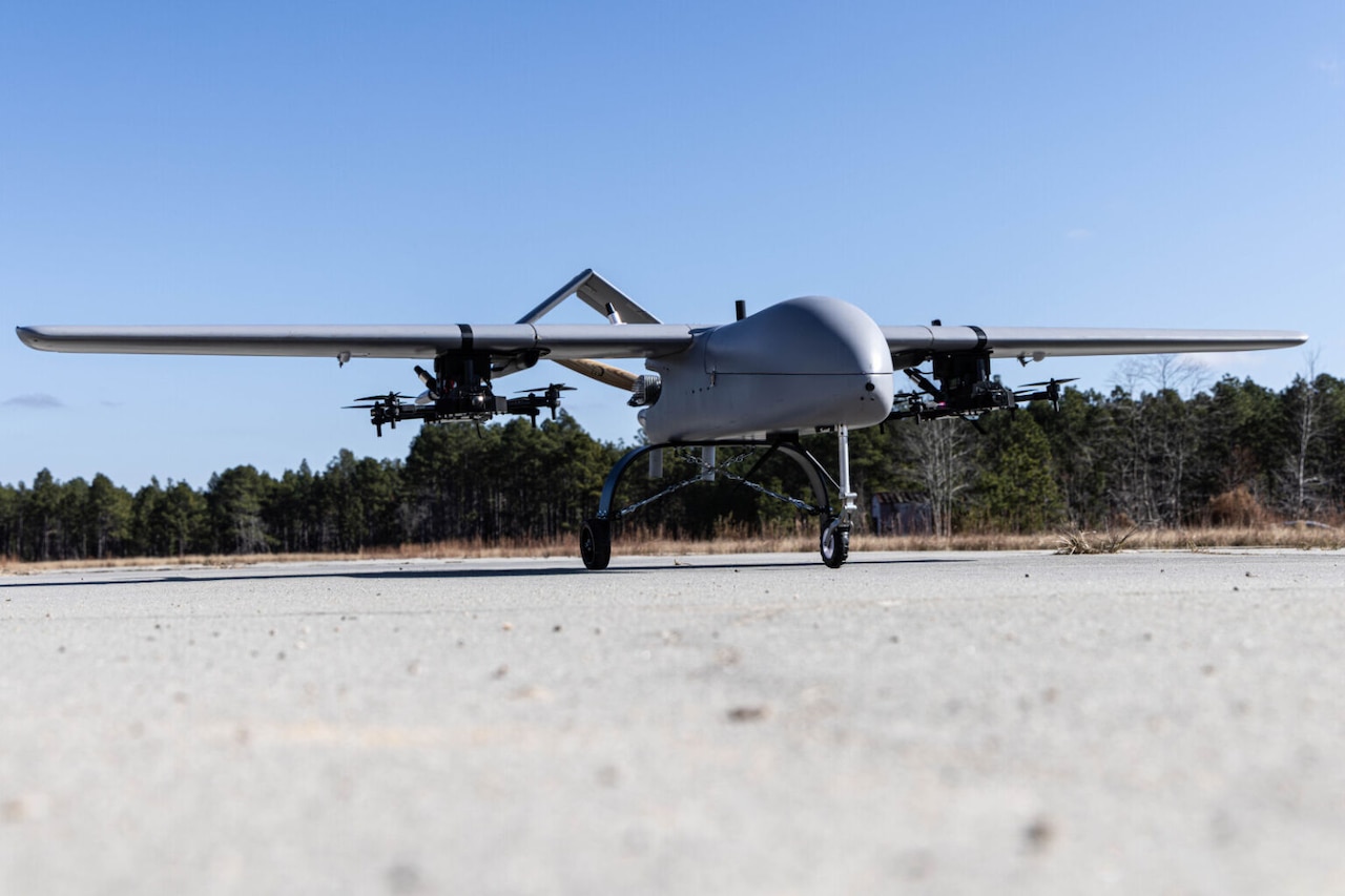 A large unmanned aerial system, with small unmanned aerial systems attached to its wings, sits on a runway.