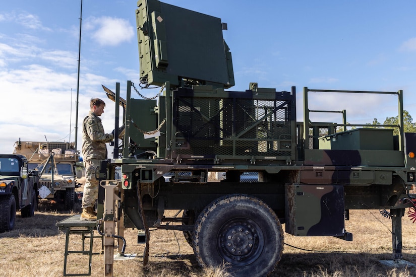 A person in a camouflage military uniform stands on a platform to work in the back of a military vehicle. Similar vehicles are in the background.