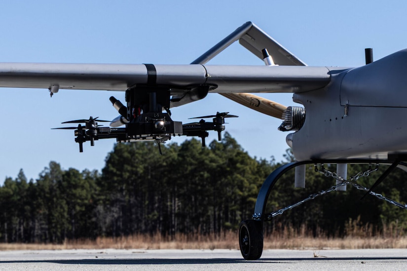 A small unmanned aerial system is attached to the bottom of the right wing of a large unmanned aerial system.