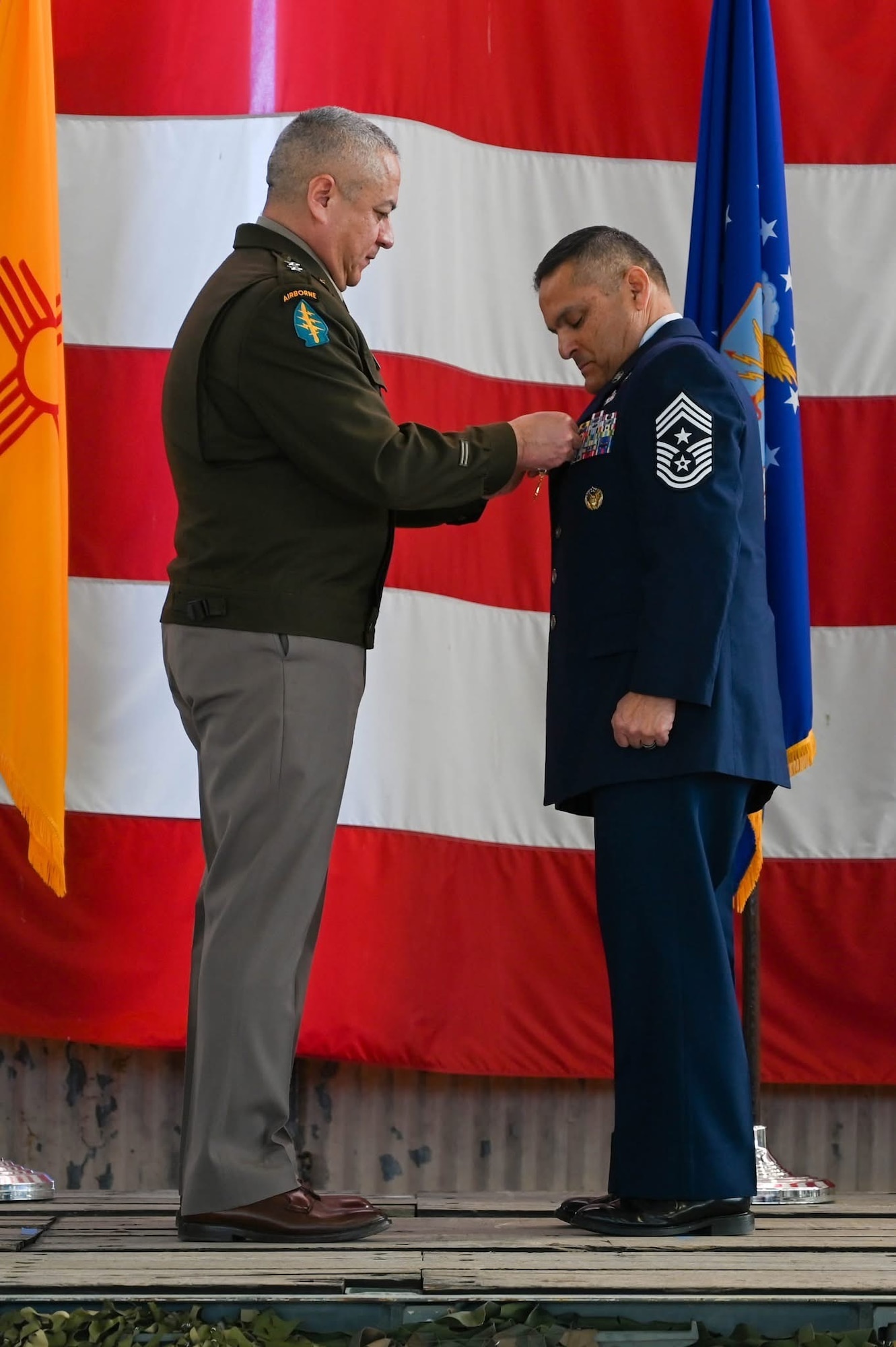 An Army general in dress uniform pins a medal on an Air Force chief in a dress uniform on a stage during a ceremony.