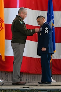 An Army general in dress uniform pins a medal on an Air Force chief in a dress uniform on a stage during a ceremony.