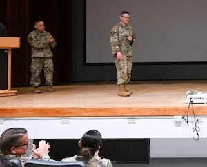 An enlisted man in an Air Force work uniform addresses a crowd from a stage.