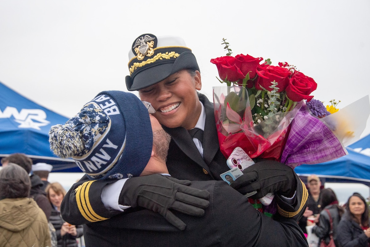 A smiling sailor holding flowers gets a kiss on the cheek from a loved one in front of a crowd in the background.
