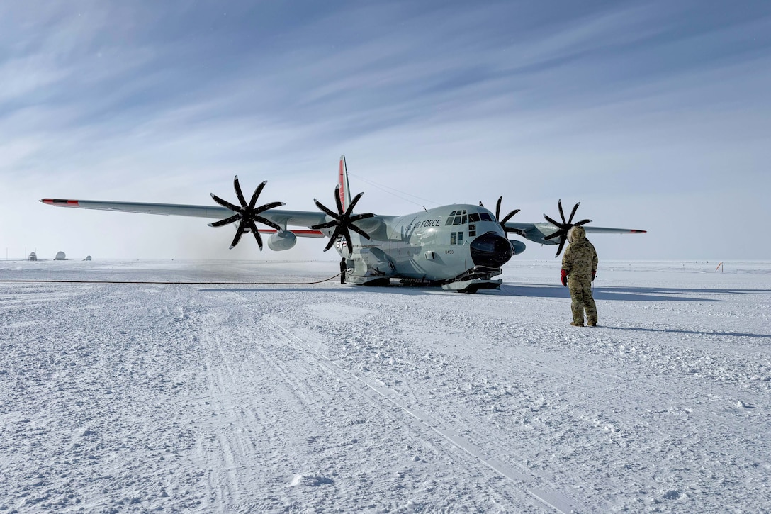 An airman fills up an aircraft with gas on a snowy tarmac as a fellow airman watches.