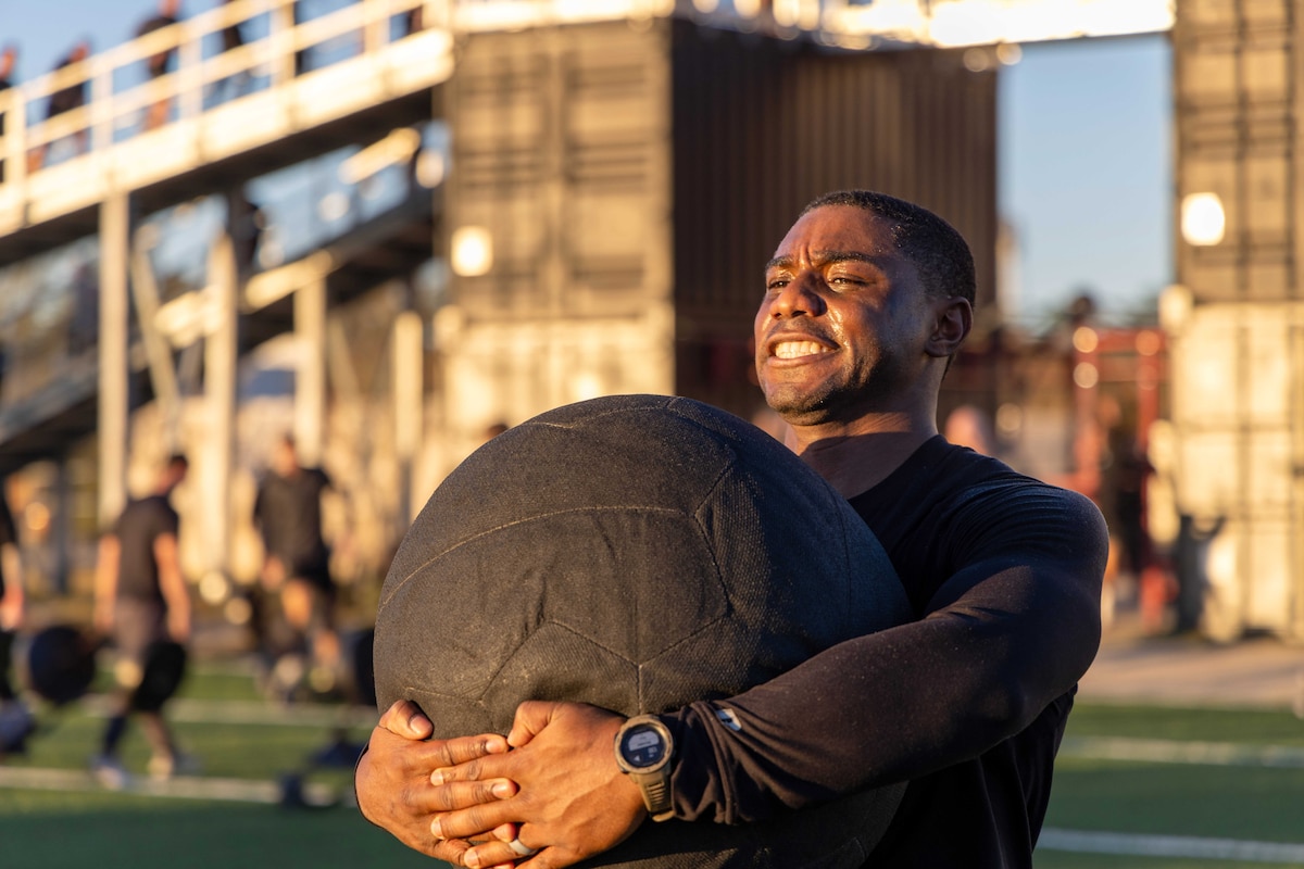 A soldier carries a weighted ball on a field as fellow soldiers train in the blurred background.