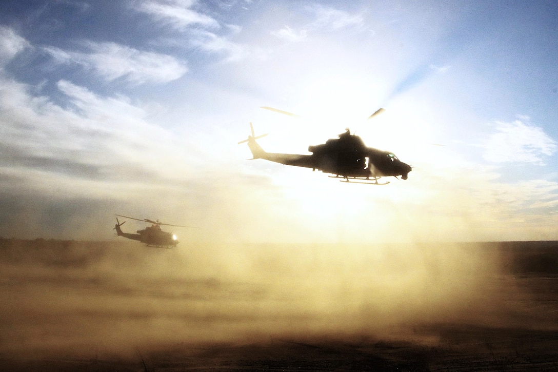 Clouds of dirt surround two helicopters as they fly over a dusty field in a sunlit sky.