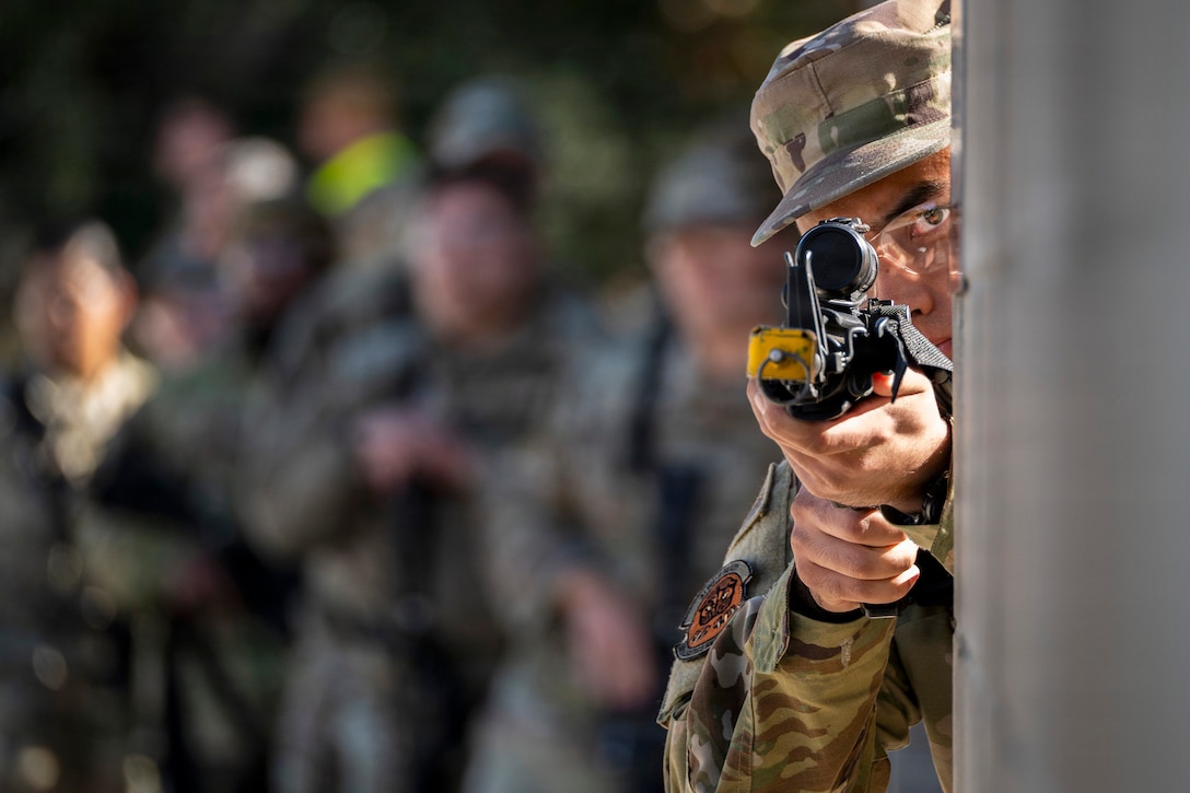 An airman peers around the corner of a building and aims a weapon.
