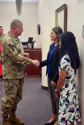 A man in Army uniform shakes the hand of a woman in a blue suit while another woman stands near.