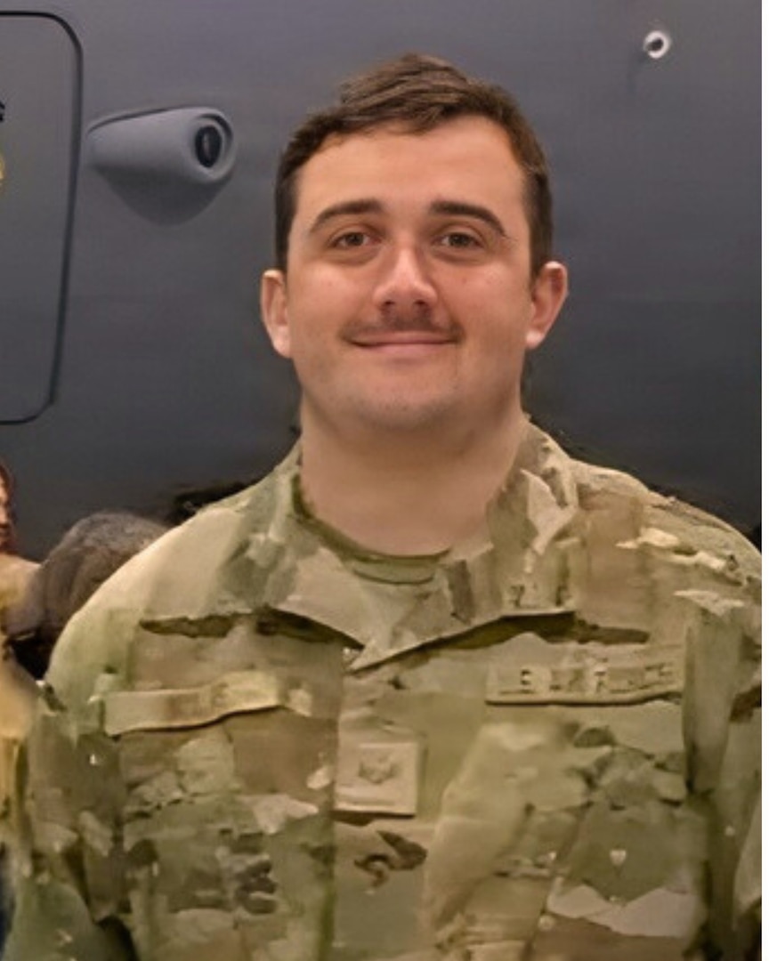 man with moustache standing in front of a grey airplane, close-up photo from mid-chest. brown hair. wearing military camo uniform.