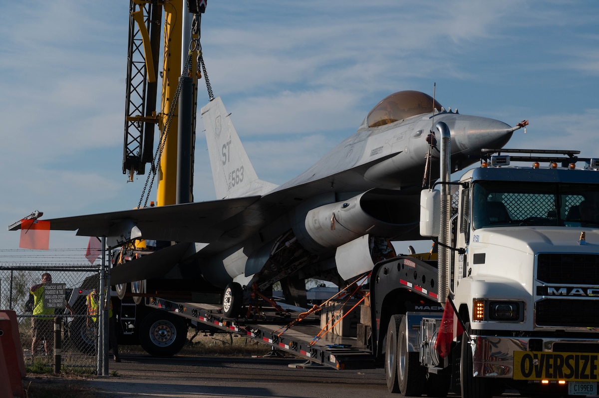 The F-16 fighter jet has arrived and is being lifted onto the back of an oversized load truck.