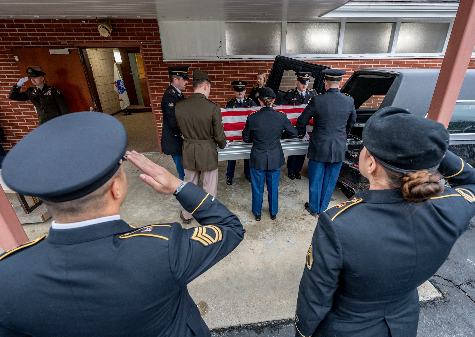 Soldiers and Armen in dress uniform saluting an American Flag-draped casket as it is loaded into a hearse.