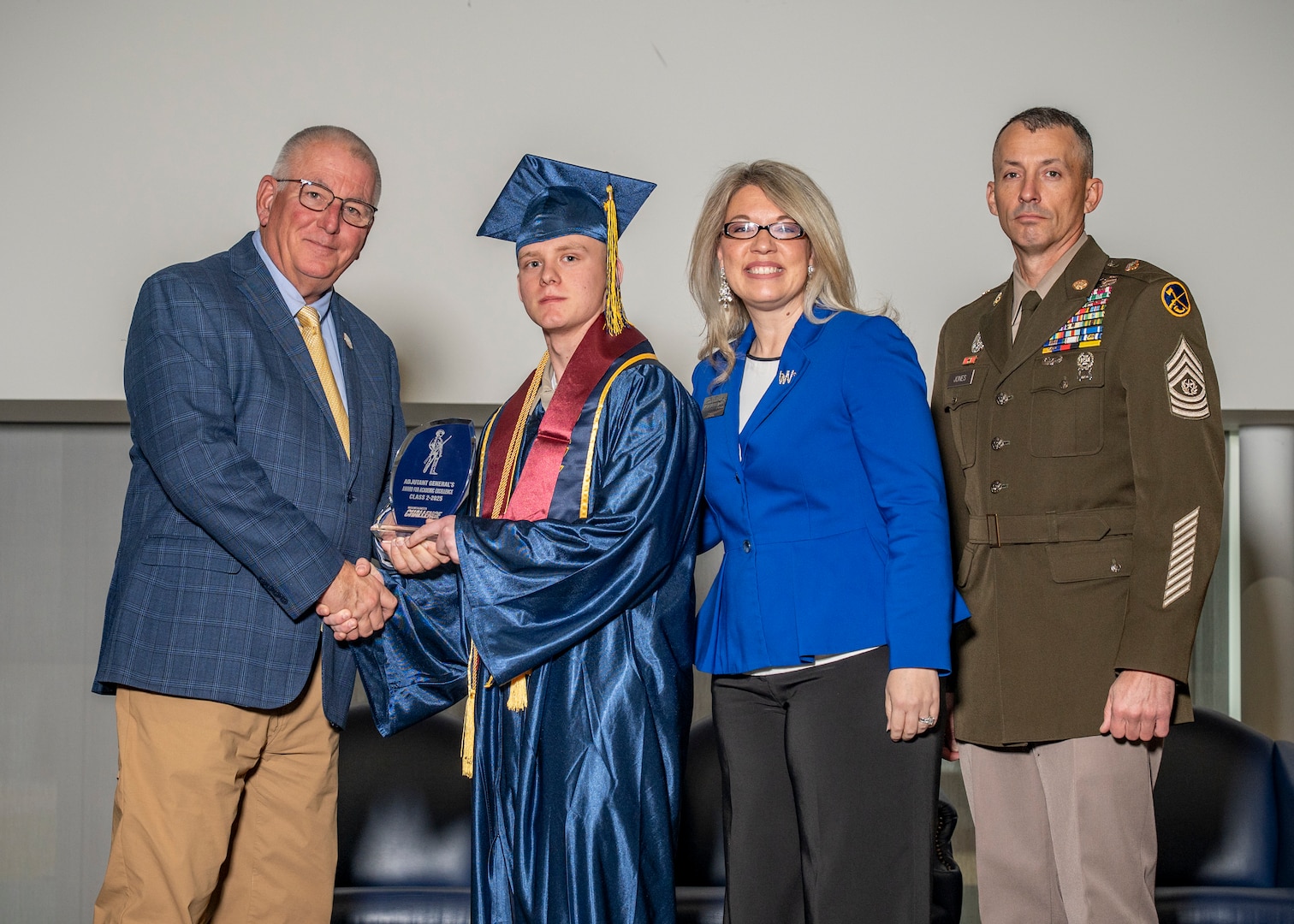 student in blue graduation robe shakes the hand of a older man in suit indoors, while a blond woman in suit and glasses and man in army military dress uniform stand at their side. all look at the camera.