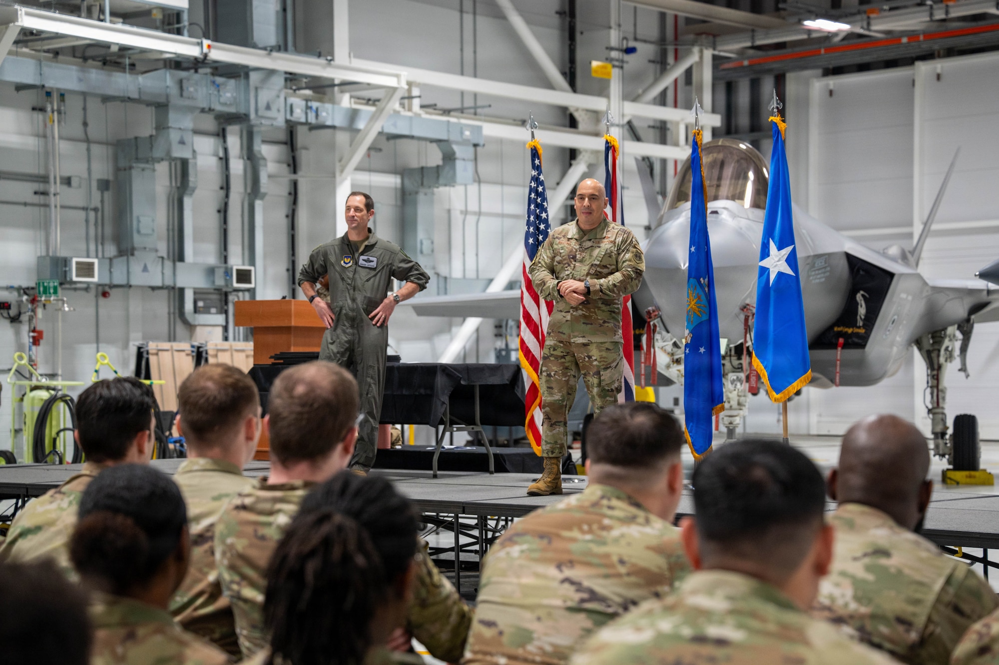 Military members listen to speakers talk on a stage.