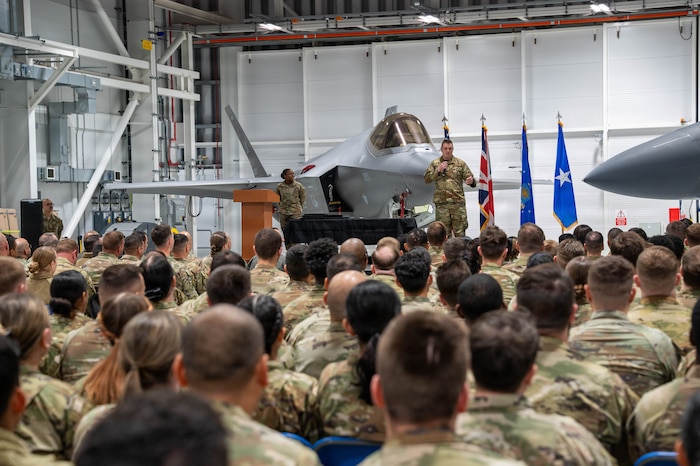 Military members listen to a speaker on a stage.
