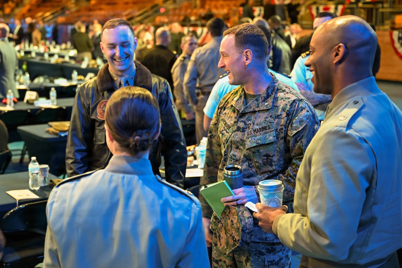 Three men and one woman wearing various military uniforms stand in a circle talking in a large auditorium. There are tables and other people talking in the background.