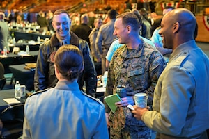 Three men and one woman wearing various military uniforms stand in a circle talking in a large auditorium. There are tables and other people talking in the background.