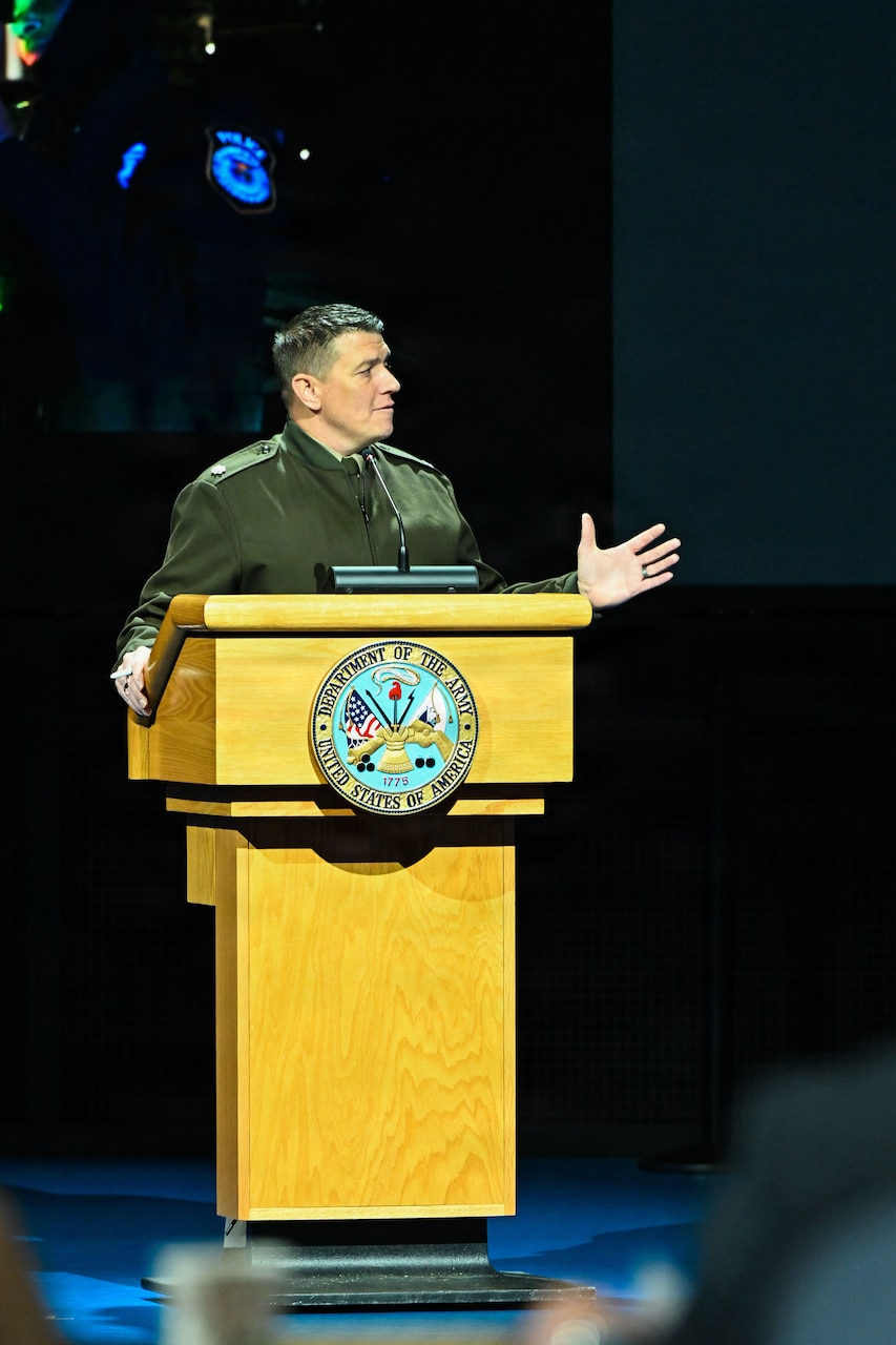 A man wearing a military dress uniform stands behind a lectern while speaking into a microphone.