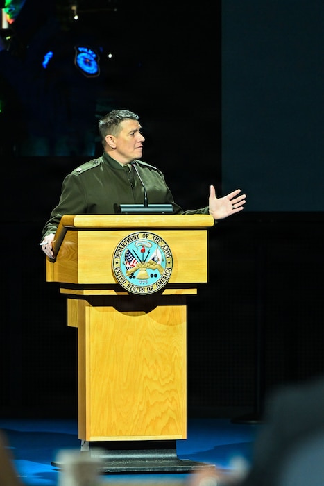 A man wearing a military dress uniform stands behind a lectern while speaking into a microphone.