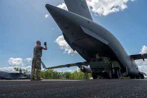 an airman in OCPs motions towards a High Mobility Artillery Rocket System entering a united states air force cargo plane