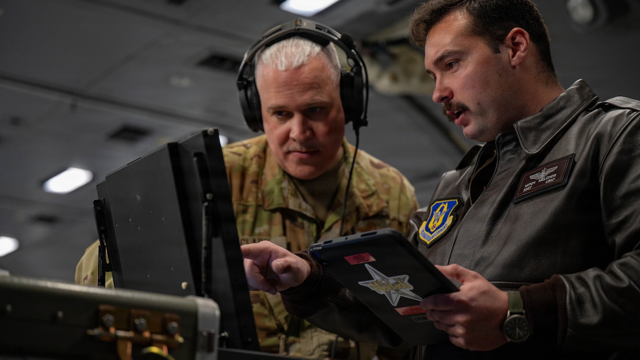 Airmen look at a tablet and laptop together aboard an aircraft